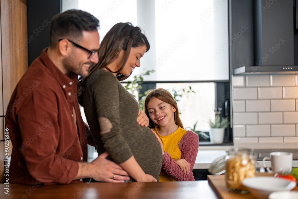 Happy little girl touching mother's pregnant belly, feeling baby at mother tummy and awaiting the birth of her little sibling. Family time in the kitchen. Happy little girl touching mother's pregnant belly, feeling baby at mother tummy and awaiting the birth of her little sibling. Family time in the kitchen.