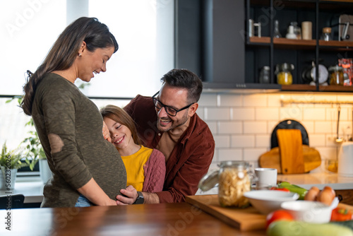 Lovely daughter and father touching mother's pregnant belly, spending some quality time together in the kitchen, being excited about the new family member. Family love and support concept.