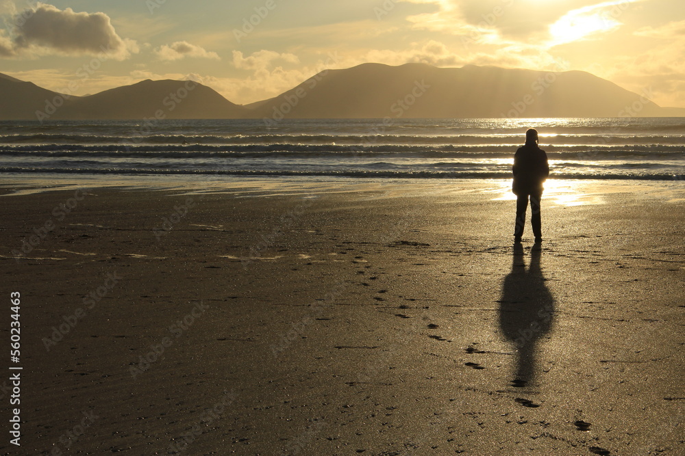 Silhouette of a person standing with their shadow behind them on the beach looking towards the mountains of Western Ireland at sunset with copy space (Inch Beach, Kerry, Ireland) Thoughtful background
