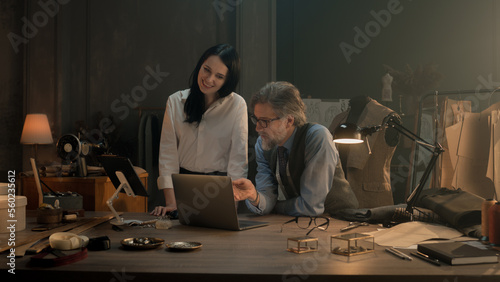 Tailors choosing design of tailored clothes collection on laptop and digital tablet computer. Colleagues working at modern workshop, talking about styles of garment, sewing type and color of cloth.