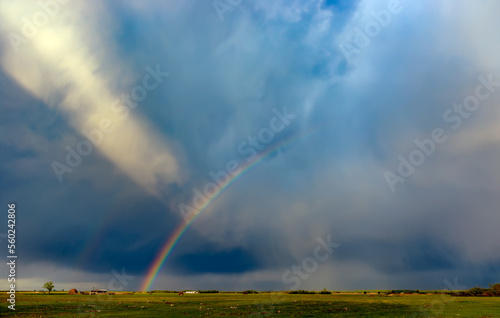 Double rainbow and a huge beam of sun light ray over dramatic stormy sky. Rural landscape with rainbow over dark stormy sky in a countryside at summer day. Scenic view of rainbow over green field