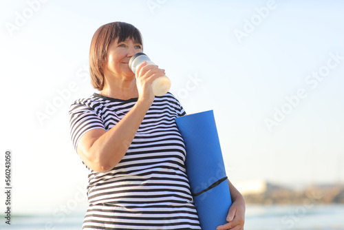mature woman drinking water from bottle after fitness exercises or yoga on beach by sea. fitness and healthy lifestyle concept - thirsty senior woman with mat maintains water balance after sport