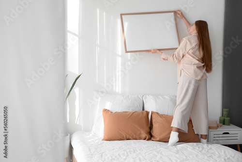 Young woman hanging blank frame on light wall in bedroom, back view photo