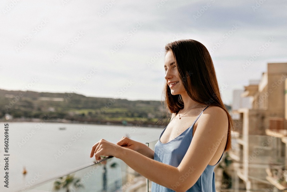 Cute charming woman with dark hair wearing blue t-shirt standing in sunny warm morning on the balcony with sea view and enjoying new day