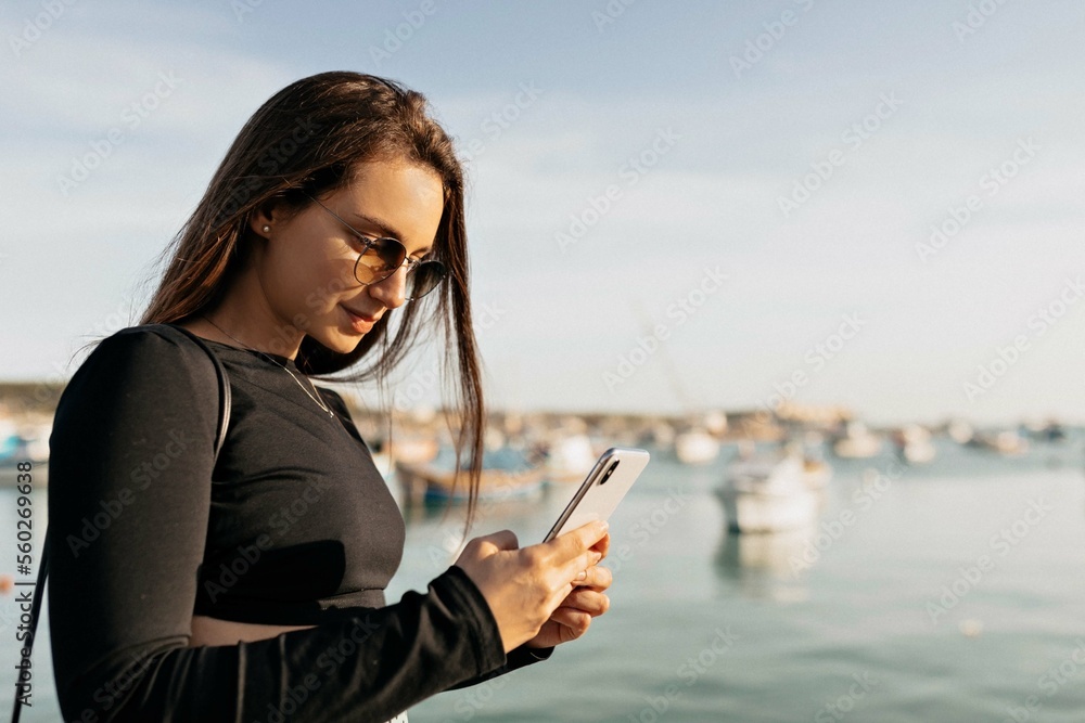 Profile outside portrait of brunette girl typing on smartphone and enjoying summer walking on background of lake in summer sunny day. Cheerful caucasian woman smiles with her teeth looking to side