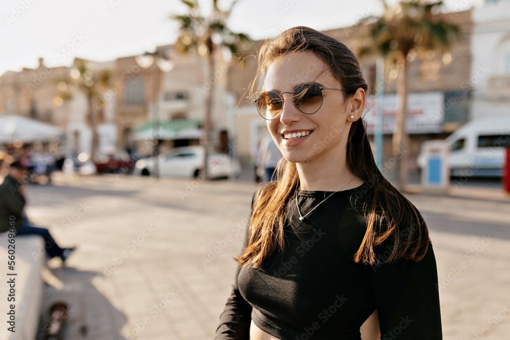 Attractive smiling happy young woman with dark hair wearing black top and sunglasses enjoying walking on quay with palms on sunset. Happy smiling European girl on vacation 