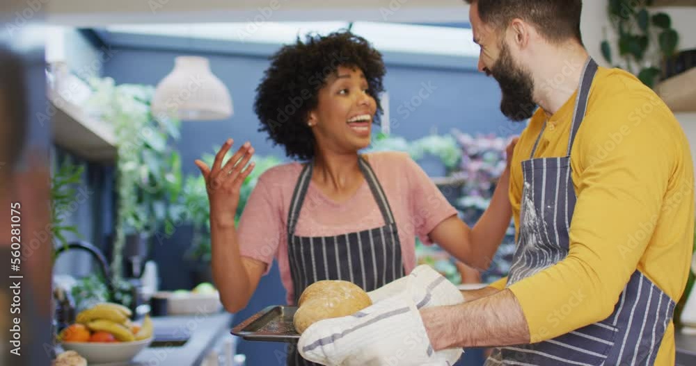 Video of happy diverse couple in aprons baking in kitchen, looking at cooked bread, with copy space Video of happy diverse couple in aprons baking in kitchen, looking at cooked bread, with copy space