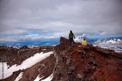 Crater of volcano. There're two people on the edge of crater of Gorely volcano. The man is standing and the woman is sitting. There're red rocks on surface. There's Mutnovskiy volcano in the backgroun photo