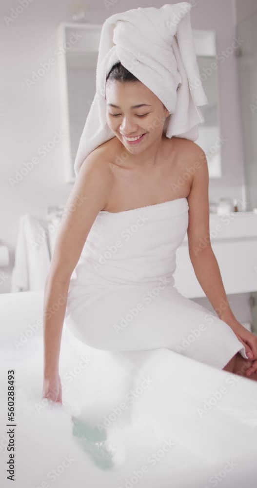Vertical video of smiling biracial woman with towel on hair sitting on bathtub in bathroom Vertical video of smiling biracial woman with towel on hair sitting on bathtub in bathroom
