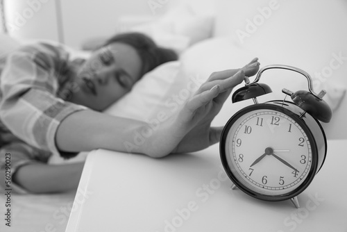 Sleepy young woman turning off alarm clock at home in morning, selective focus. Black and white photography