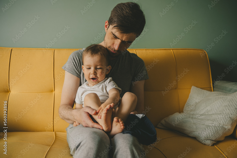 Father dresses infant in living room sitting on couch at home. Clothes baby undershirts and sliders on baby close-up. Home care and baby care concept. Father dresses infant in living room sitting on couch at home. Clothes baby undershirts and sliders on baby close-up. Home care and baby care concept.