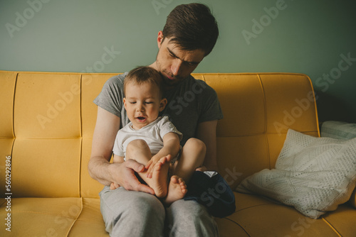 Father dresses infant in living room sitting on couch at home. Clothes baby undershirts and sliders on baby close-up. Home care and baby care concept.