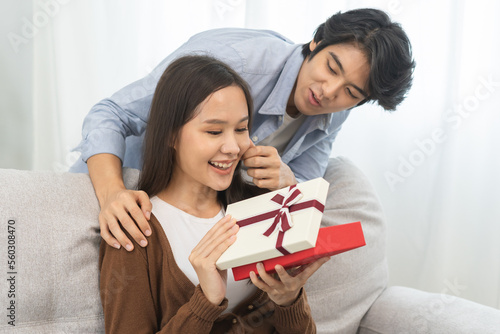 Celebrating Valentine's day anniversary, relationship two asian young couple love, boyfriend gives a gift to girlfriend by hide box at the behind, woman getting present while sitting on sofa at home.