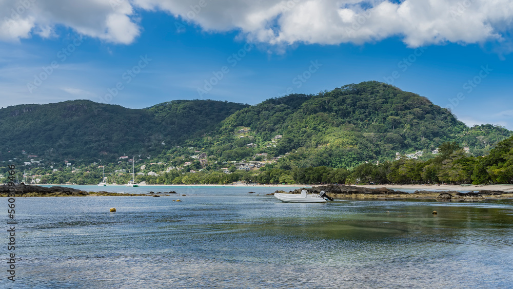 Low tide on a tropical island. The white boat is moored in shallow water. The stones of the exposed seabed are visible. Green hills against a blue sky of clouds. Seychelles. Mahe. Beau Vallon