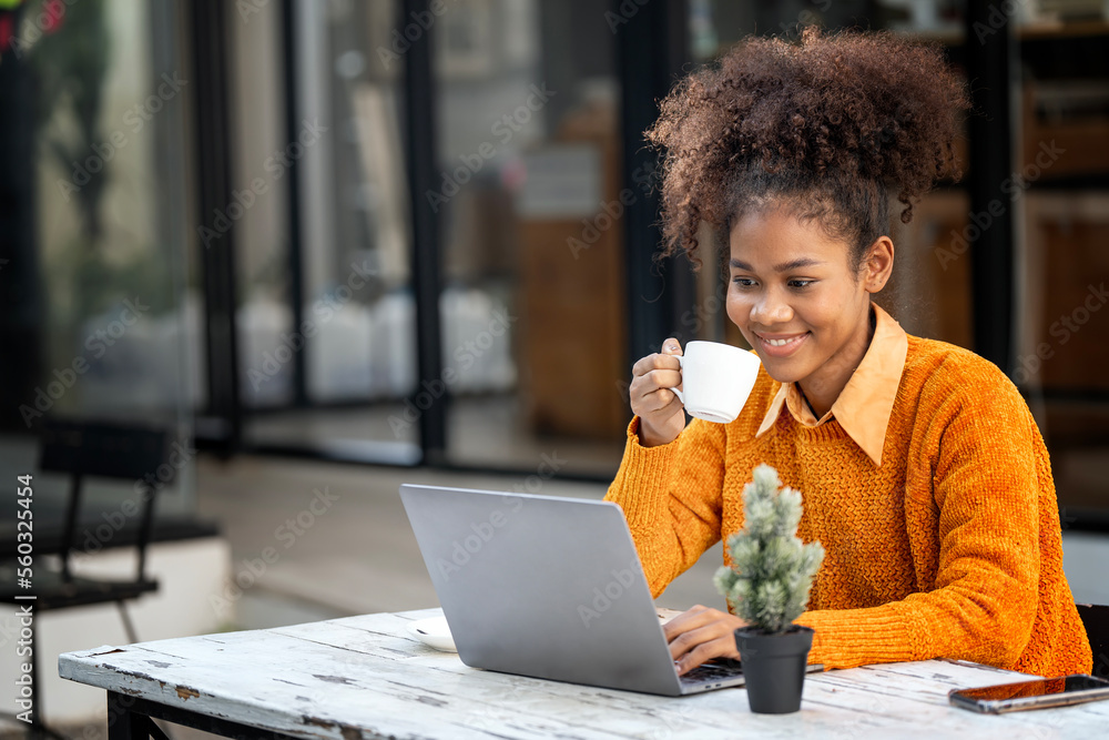 Attractive african young confident woman holding coffee cup and using laptop computer, sitting at open co-workspace. - obrazy, fototapety, plakaty 