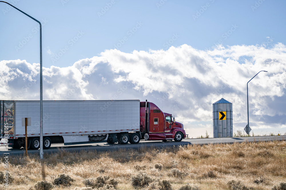 Dark red big rig classic American semi truck tractor transporting cargo in refrigerator semi trailer going uphill on the local road at sunny day