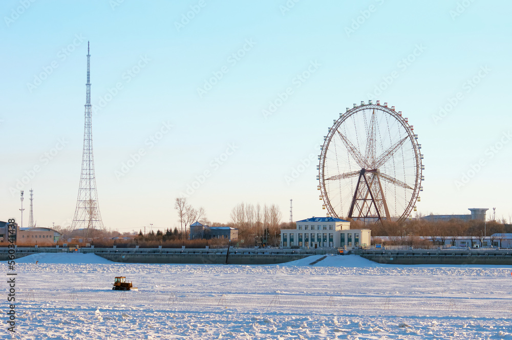 Ferris wheel on the bank of a snowy river against the backdrop of a beautiful sky. Tractor rides on ice on the state border. Winter landscape.