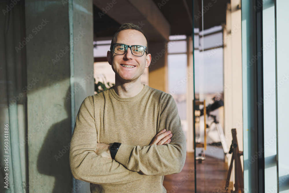 A young man in glasses and casual clothes stands in a modern workspace. Male freelancer works remotely. A young man in glasses and casual clothes stands in a modern workspace. Male freelancer works remotely.