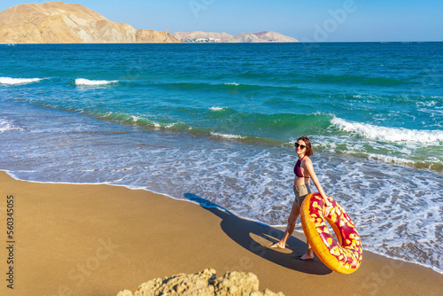 Teen girl with donut lilo against summer landscape of sea and mountains on sunny day. Beautiful young woman in swimwear and sunglasses with inflatable ring on beach. Vacation, happy lifestyle concept