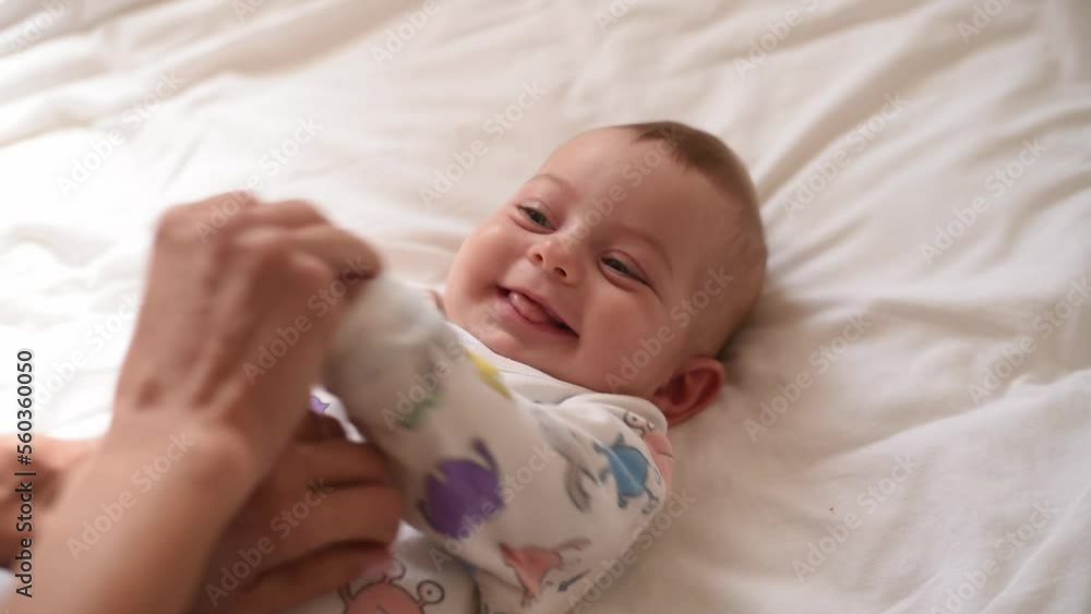 Smiling baby in white t-shirt with colorful application lies on back on white bed. Close-up. Mother's hands help move child's hands. First year of life and knowledge of world around. Side view Smiling baby in white t-shirt with colorful application lies on back on white bed. Close-up. Mother's hands help move child's hands. First year of life and knowledge of world around. Side view