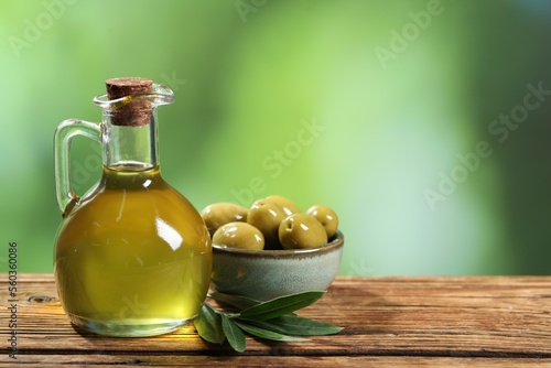 Jug of cooking oil, olives and green leaves on wooden table against blurred background. Space for text