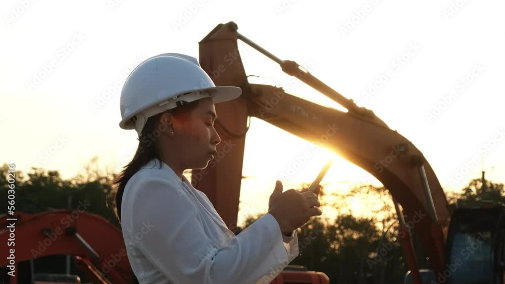 Portrait of an Asian engineer wearing a white helmet using a smartphone in a workplace. Civil engineer checking after work on construction site during sunset on yellow excavator background. Portrait of an Asian engineer wearing a white helmet using a smartphone in a workplace. Civil engineer checking after work on construction site during sunset on yellow excavator background.