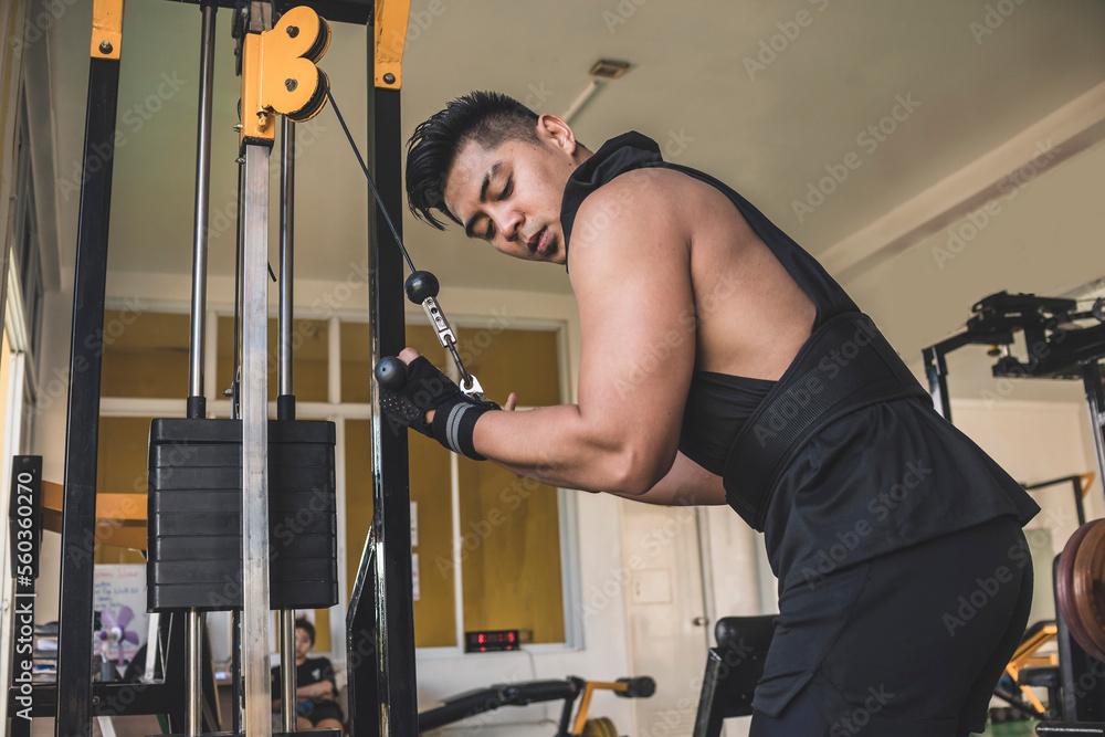 A serious and tanned asian guy doing reverse grip cable tricep push downs using a straight pushdown bar. Feeling the squeeze in his arms. Wearing a black hooded low-cut tank top. A serious and tanned asian guy doing reverse grip cable tricep push downs using a straight pushdown bar. Feeling the squeeze in his arms. Wearing a black hooded low-cut tank top.