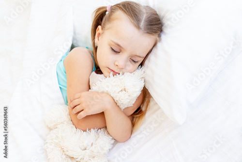 a girl child is sleeping on a bed at home on a white cotton bed hugging a teddy bear toy in her hands and smiling sweetly in her sleep