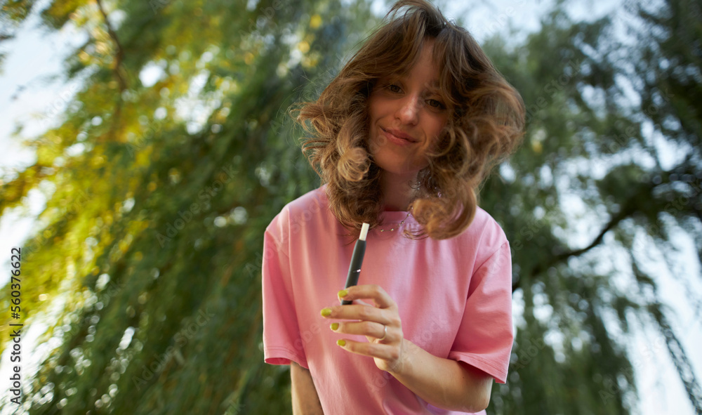 Cheerful girl in a pink t-shirt with an electronic cigarette in the park. Beautiful girl in a pink t-shirt, beaming smile, holding an electronic cigarette. Teenage girl using an electronic cigarette. Cheerful girl in a pink t-shirt with an electronic cigarette in the park. Beautiful girl in a pink t-shirt, beaming smile, holding an electronic cigarette. Teenage girl using an electronic cigarette.
