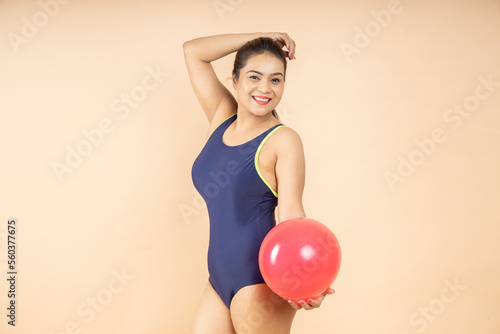 Happy young brunette asian woman slim body wearing blue one-piece swimsuit hold play red beach ball isolated on beige background, studio shot. Summer holidays and travel concept. © gajendra