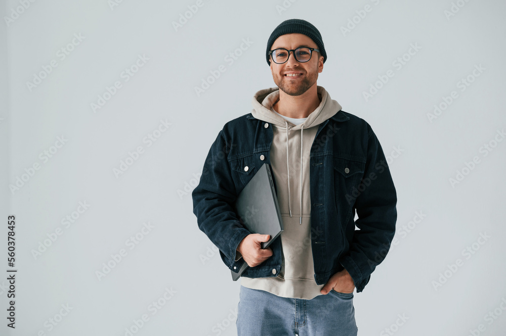 IT specialist in hat and glasses standing with laptop. Handsome man is in the studio against white background IT specialist in hat and glasses standing with laptop. Handsome man is in the studio against white background