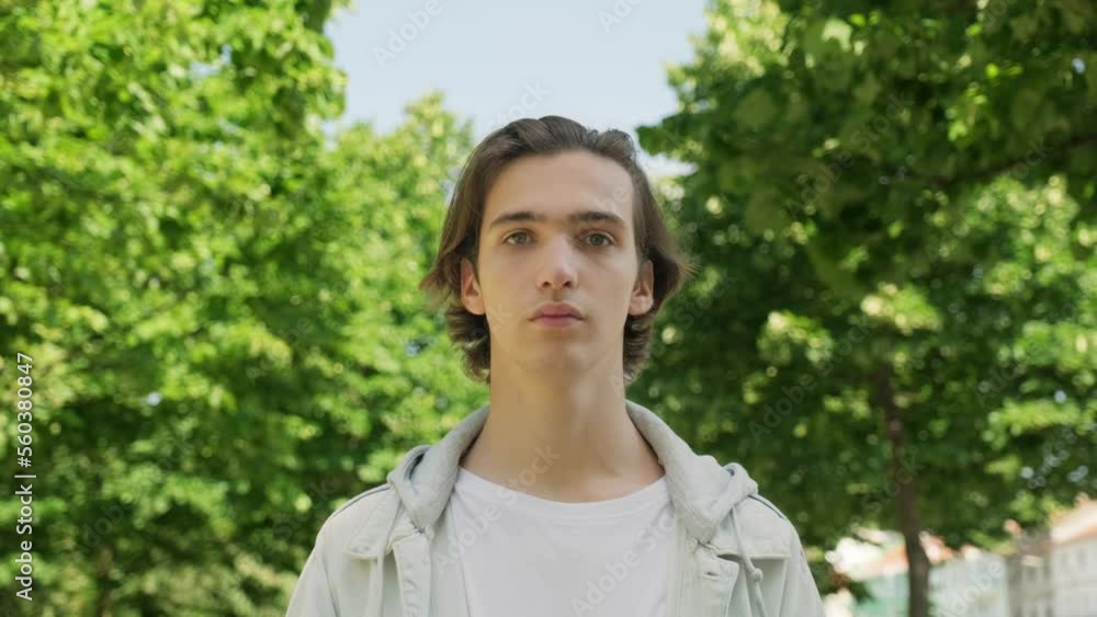 Serious young man stands motionless and looks in the camera, outdoors. Camera zoom in to the Calm and peaceful face of an young man. Closeup calm face of handsome guy at the park. Slow motion Serious young man stands motionless and looks in the camera, outdoors. Camera zoom in to the Calm and peaceful face of an young man. Closeup calm face of handsome guy at the park. Slow motion