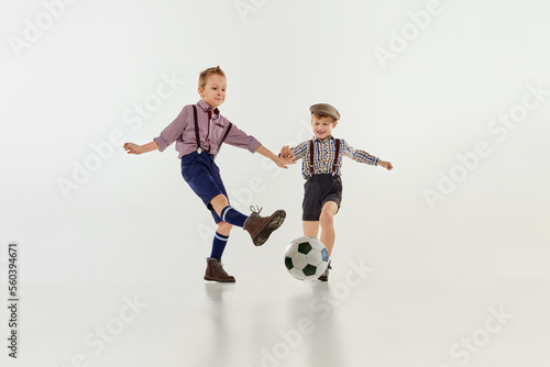 Competition. Boys, children in classical retro clothes playing football over grey studio background. Concept of game, childhood, friendship