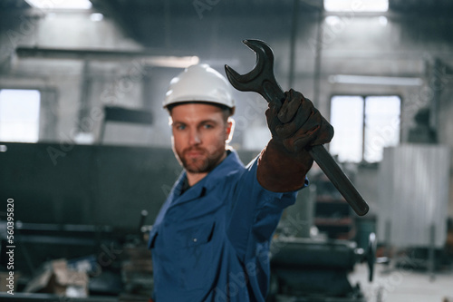 Wrench in the hand. Factory worker in blue uniform is indoors