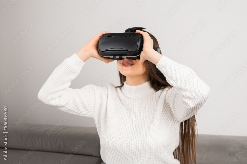 Woman puts on VR goggles to immerse into virtual reality. Young housewife sits on comfortable sofa using modern technology at home Woman puts on VR goggles to immerse into virtual reality. Young housewife sits on comfortable sofa using modern technology at home