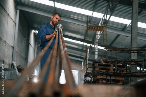 With bunch of metal pipes. Factory worker in blue uniform is indoors