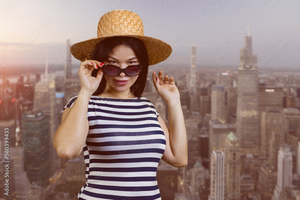 Portrait of attractive young asian woman in sunglasses and straw hat. Big cityscape background.