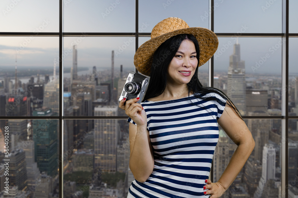 Happy young tourist asian woman is holding a vintage photo camera. Checkered windows with cityscape view on background.