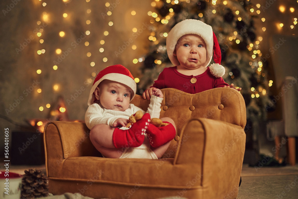 Baby is on the little sofa. Two kids in Santa hats are celebrating holidays indoors together