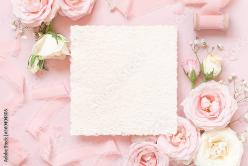 Blank square card between pink roses and pink silk ribbons on pink top view, wedding mockup