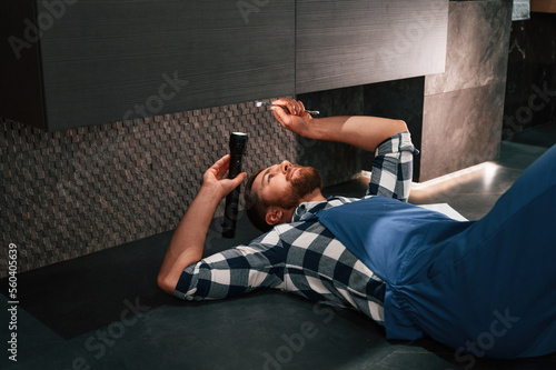 Fixing the siphone. Lying down on the floor. Plumber in blue uniform is at work in the bathroom