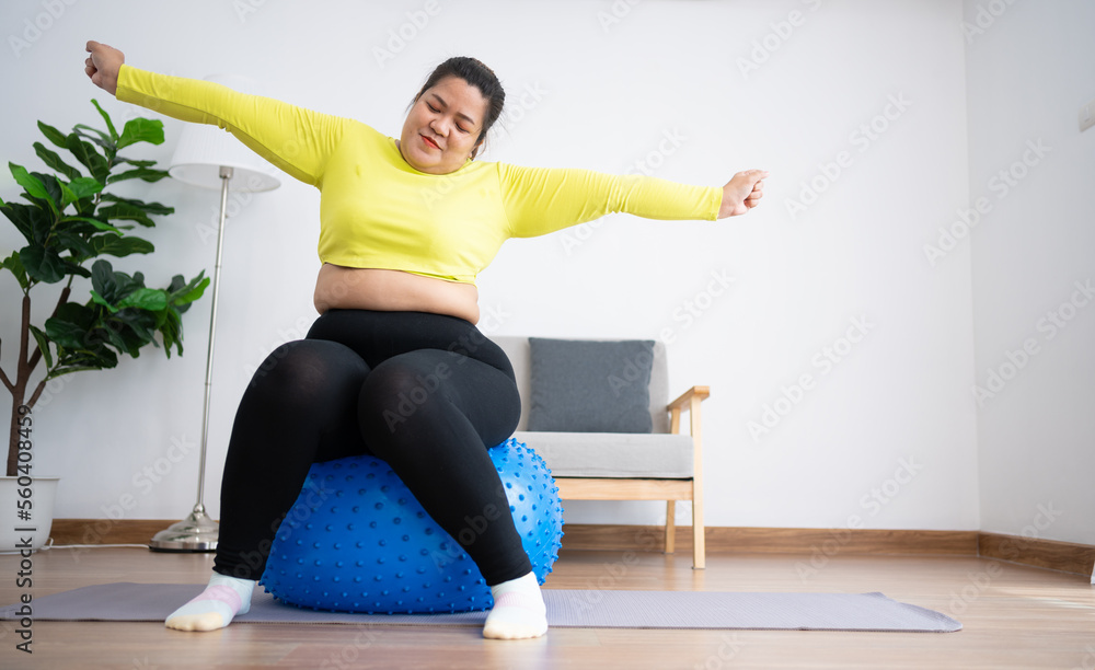 Portrait plus size woman doing exercise with fitness ball in home gym. Overweight woman sitting on a pilates ball and Stretching her muscles before exercising. Health care and weight loss concept. Portrait plus size woman doing exercise with fitness ball in home gym. Overweight woman sitting on a pilates ball and Stretching her muscles before exercising. Health care and weight loss concept.
