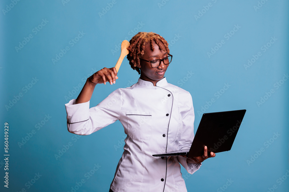 African american smiling woman, uniformed professional chef, searching on internet with computer, vertical studio portrait. Confused cooker looking dish preparing instructions on laptop screen.