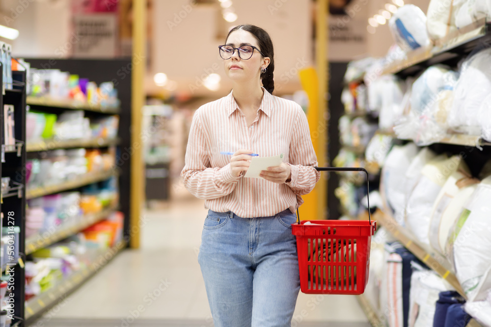 Young Caucasian woman wearing glasses holds grocery basket, holds list and selects bed linen and pillow. Shopping and purchase concept