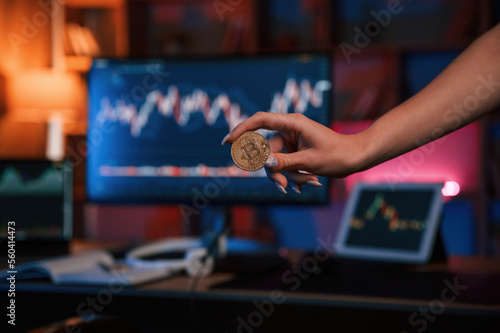 Close up view of woman's hand that holding bitcoin against computer display and tablet with graphs