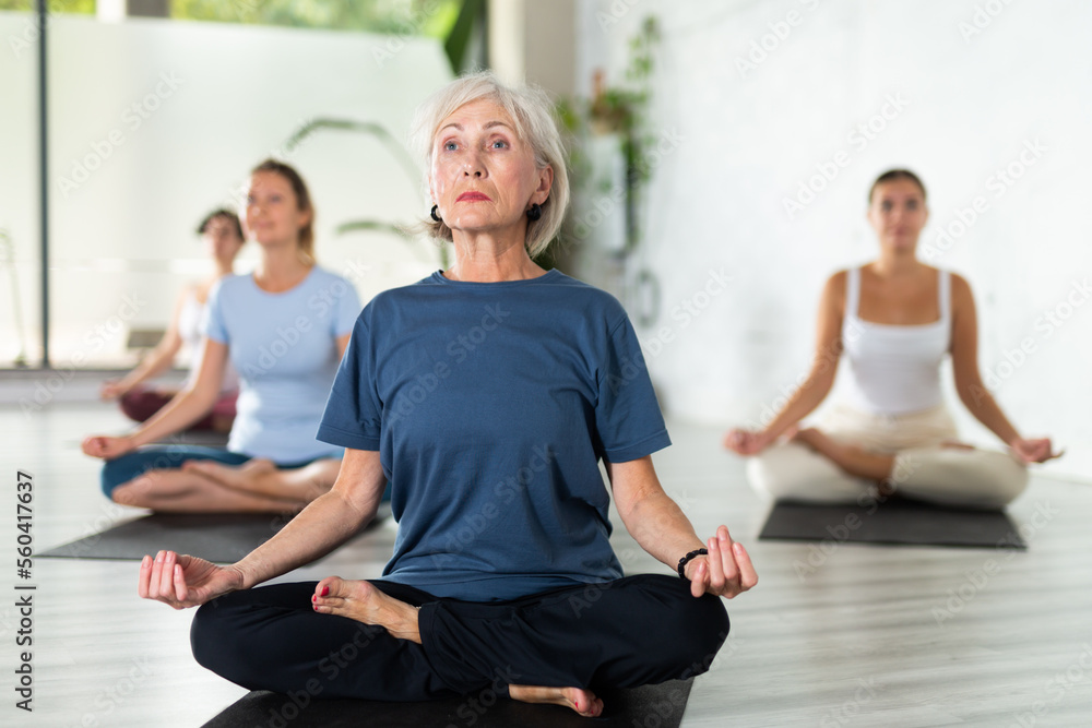 Peaceful mature female sitting in Lotus Pose Padmasana on mat during group yoga class in fitness studio