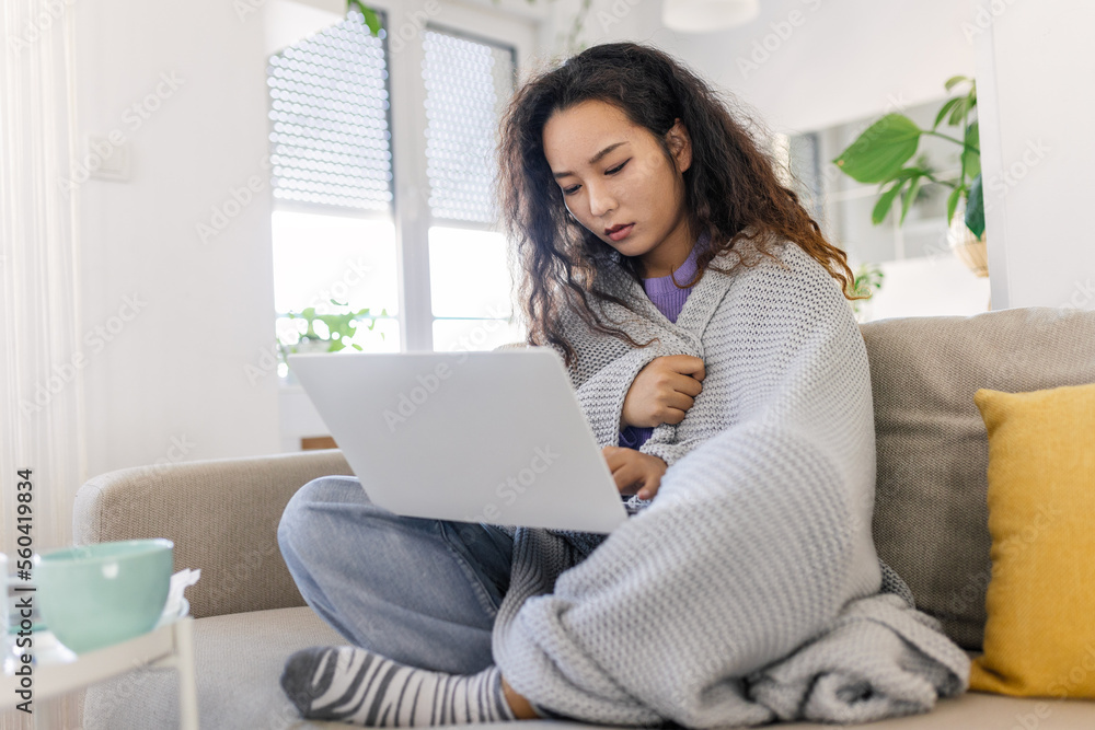 Asian woman wrapped in a cozy blanket sitting on the sofa and working on her laptop Asian woman wrapped in a cozy blanket sitting on the sofa and working on her laptop