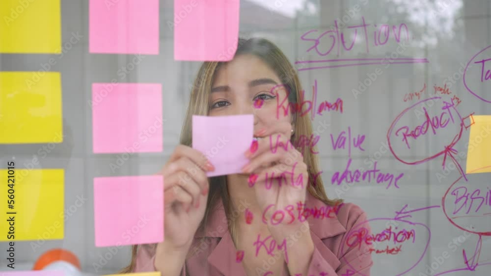 Young Creative businesswoman holding a marker and writing plan and share idea on glass wall with sticky note, Brainstorming and discussing and formulating, business strategies in tech startup office. Young Creative businesswoman holding a marker and writing plan and share idea on glass wall with sticky note, Brainstorming and discussing and formulating, business strategies in tech startup office.