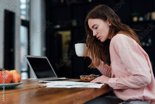 Young woman works with her laptop while eating breakfast in the kitchen