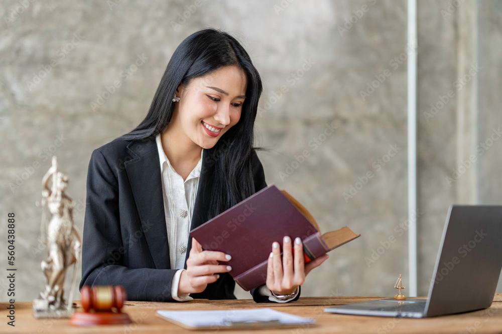 Obraz premium Asian female lawyer or legal advisor working with scales of justice Sitting at her desk holding a book on the scale of jurisprudence concepts to study and find information.